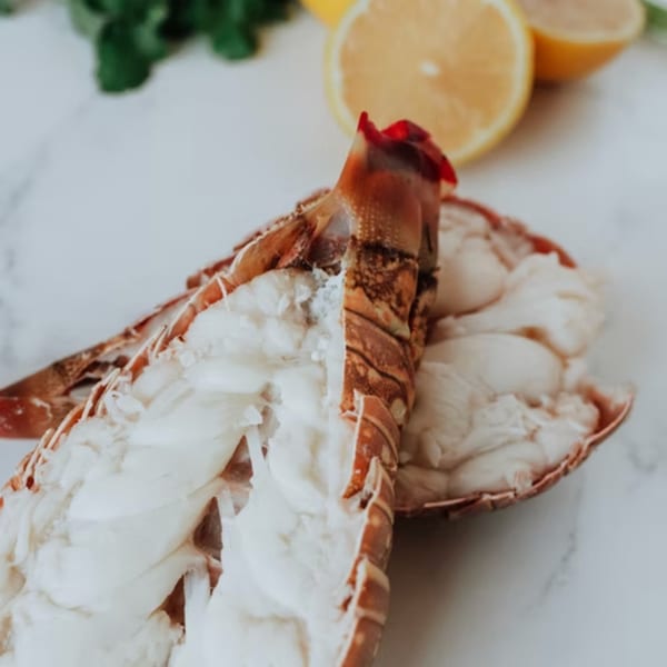 Two raw lobster tails with lemons and herbs in the background on a white marble surface.